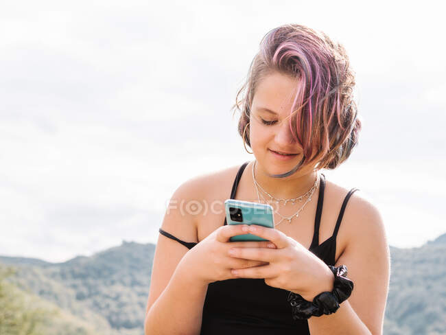 Позитивна жінка з пофарбованими повідомленнями про волосся на сучасному мобільному телефоні, проводячи час у природі з гірським хребтом у високогір'ї — стокове фото