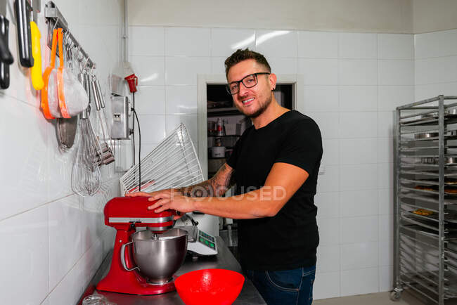Positive young unshaven male confectioner in casual clothes and eyeglasses smiling and looking at camera while using modern stand mixer during pastry preparation in bakery kitchen — Stock Photo