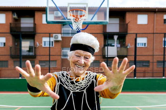 Улыбающаяся взрослая женщина в спортивной одежде и белой сетке, смотрящая в камеру, стоя на спортивной площадке с баскетбольным обручем во время тренировки — стоковое фото
