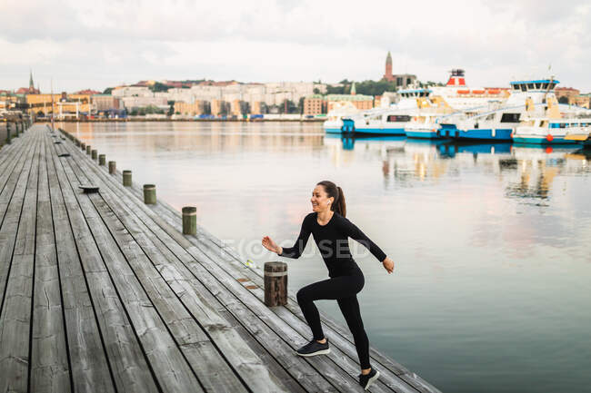 Vista laterale del corpo pieno di femmina sportiva in activewear che corre su argine di legno vicino al fiume calmo con barche durante l'allenamento — Foto stock