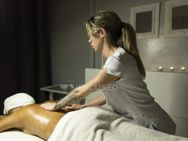 Masajista femenina irreconocible masajeando la espalda del cliente en toalla en el sofá en el centro de spa ligero - foto de stock