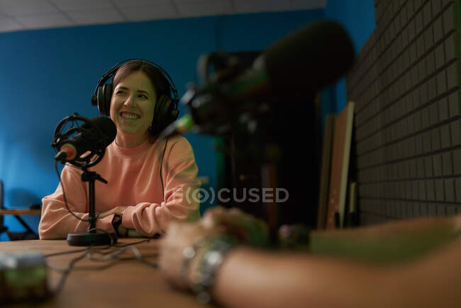 Heureux animateur de radio féminine en vêtements décontractés et écouteurs souriant tout en enregistrant podcast en studio moderne avec la récolte collègue méconnaissable — Photo de stock