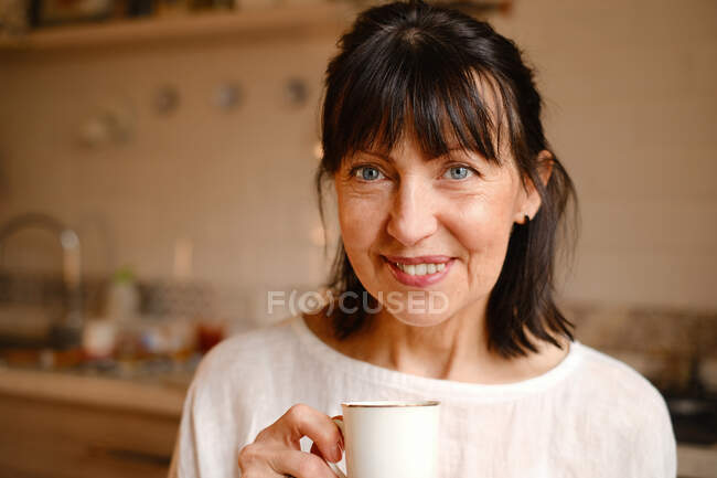 Позитивна жінка середнього віку яскраво посміхається, насолоджуючись гарячим напоєм на кухні вранці і дивлячись на камеру — стокове фото