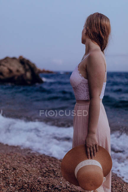 Зачарована жінка з довгим волоссям у модному платті, що стоїть на березі моря влітку в той час як вона тримає капелюх однією рукою — стокове фото