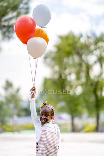 Allegro ragazza afroamericana con trecce in abiti eleganti in esecuzione con palloncini colorati in mano nel parco durante il giorno — Foto stock