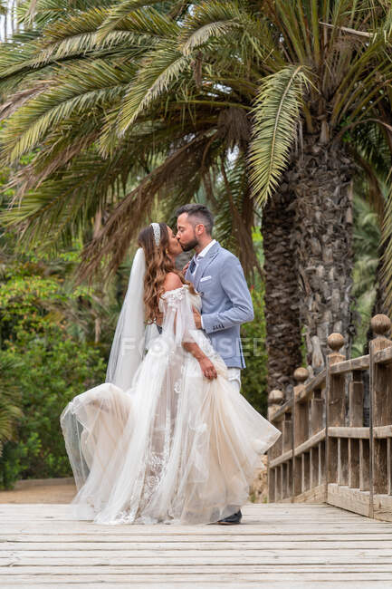 Супружеская пара в свадебных нарядах, стоящая на деревянном пешеходном мосту с перилами, целуясь возле зеленых пальм и растений в саду в летний день — стоковое фото
