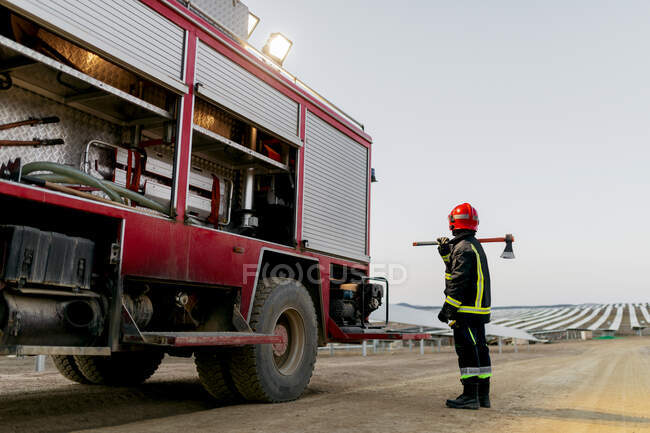 Full length of brave anonymous firefighter wearing special protective uniform and holding axe on shoulder while standing near red fire truck and looking away in countryside — Stock Photo