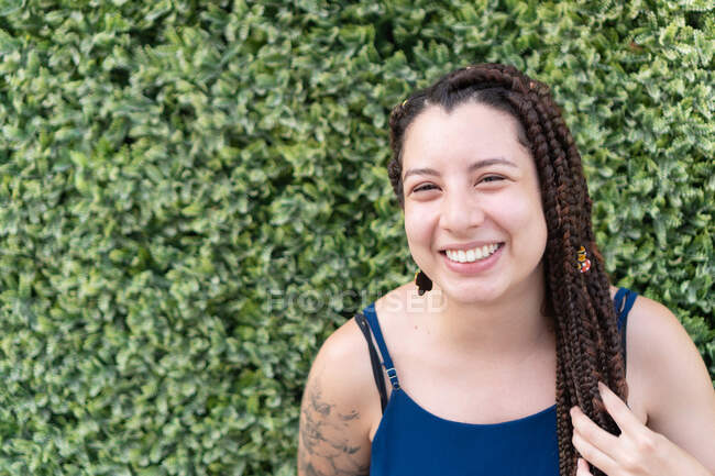 Позитивная латиноамериканка с татуировкой и длинными черными плетеными волосами улыбается и смотрит в камеру, стоя рядом с зелеными растениями — стоковое фото