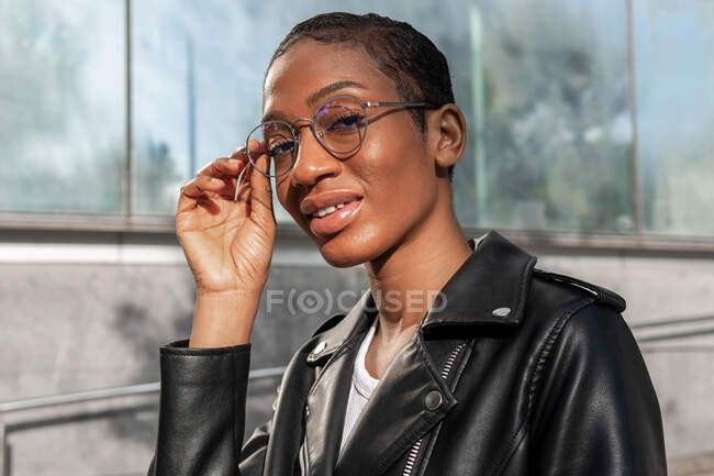 Jolie femme afro-américaine aux cheveux courts en veste en cuir ajustant les lunettes à la mode et regardant la caméra sur la rue ensoleillée près du bâtiment — Photo de stock