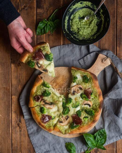 Верхний вид на неузнаваемый урожай, принимающий кусочек вкусной пиццы с соусом песто и грибами, подаваемый на деревянной доске для резки на столе — стоковое фото