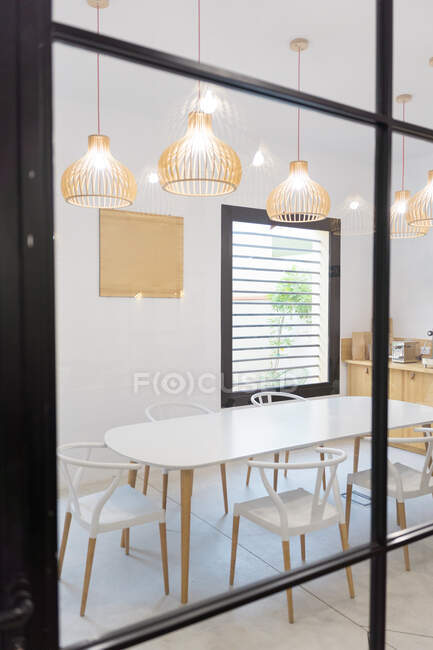 A través de vidrio de sillas blancas y mesa colocada cerca de mostrador y ventana en la cafetería de luz moderna con lámparas brillantes colgantes - foto de stock