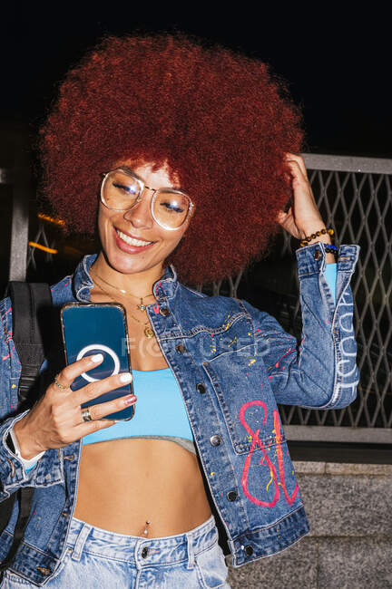 Mujer alegre con peinado afro que usa ropa de mezclilla de moda mensajes de texto en el teléfono celular mientras está cerca de la valla en la calle por la noche - foto de stock