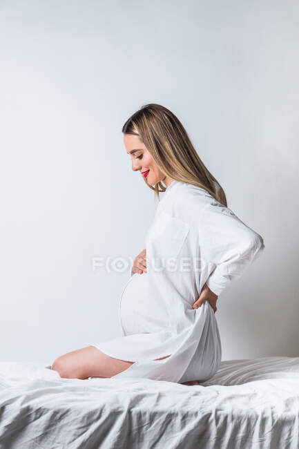 Vue latérale de la jeune femelle douce touchant le ventre tout en étant assis sur le lit et souriant joyeusement — Photo de stock