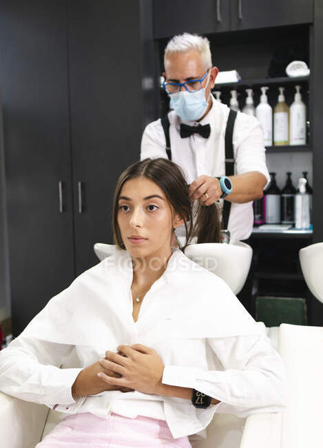 Grauhaariger Friseur mit medizinischer Maske und Brille wäscht Haare einer Frau in modernem Salon — Stockfoto