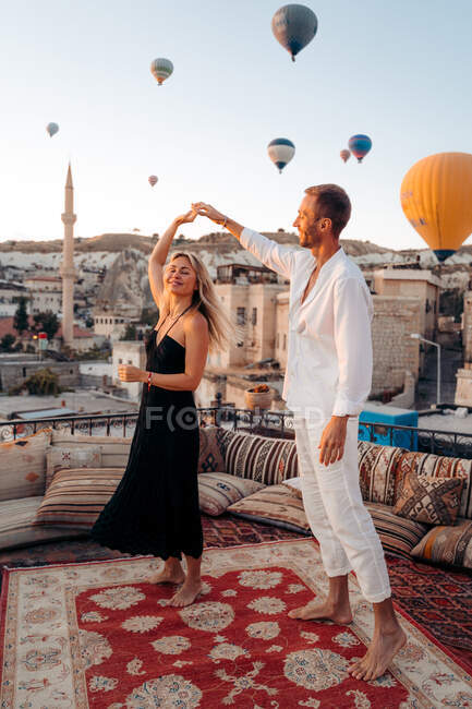 Full body of barefoot couple dancing together on rooftop terrace against hot air balloons flying in cloudless sky — Stock Photo