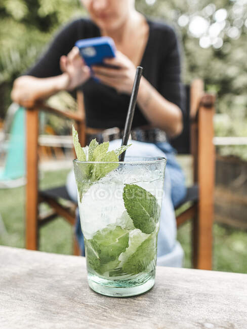 Bicchiere di cocktail rinfrescante con ghiaccio e foglie di menta posto su tavolo di legno su sfondo sfocato di donna che naviga smartphone nel giardino estivo — Foto stock