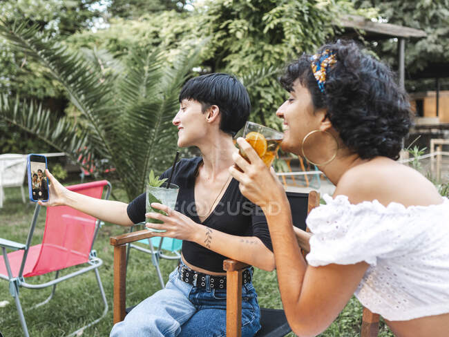 Délicieuse multiraciale meilleures amies avec des verres de cocktails se refroidissant dans le parc d'été et prenant selfie sur smartphone — Photo de stock