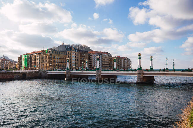 Pintoresco paisaje urbano con edificios residenciales situados cerca del canal del río con aguas tranquilas bajo el cielo azul nublado en Donostia en España - foto de stock