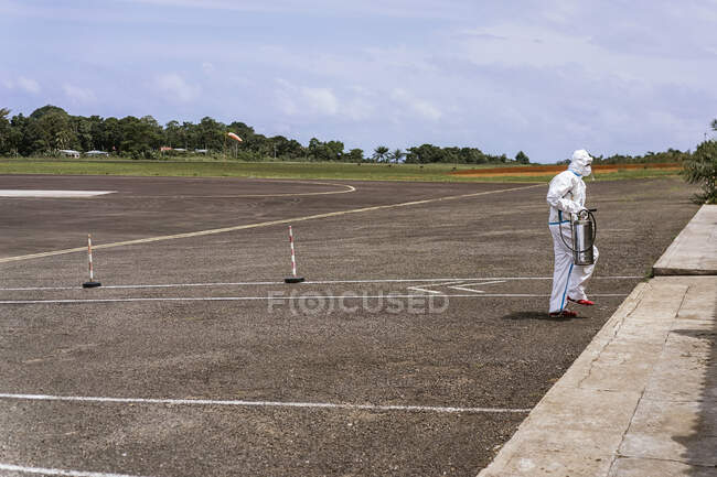 Vista lateral de médico irreconocible en uniforme protector blanco caminando en camino de asfalto vacío en So Tom y Prncipe durante la pandemia de coronavirus - foto de stock