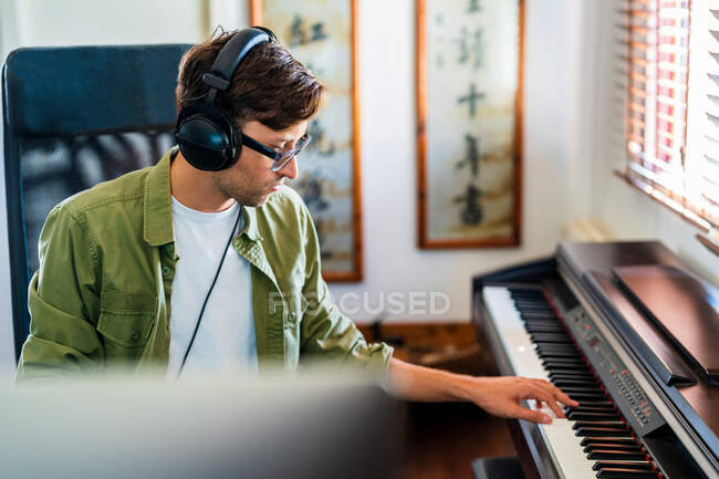 Вид сбоку мужчины-музыканта в повседневной одежде, играющего на фортепиано, сидя в современной квартире у окна — стоковое фото