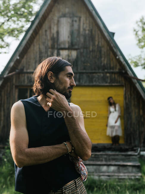 Close-up of a serious man looking away and in the background an blurred girl leaning against an abandoned wooden house — Stock Photo