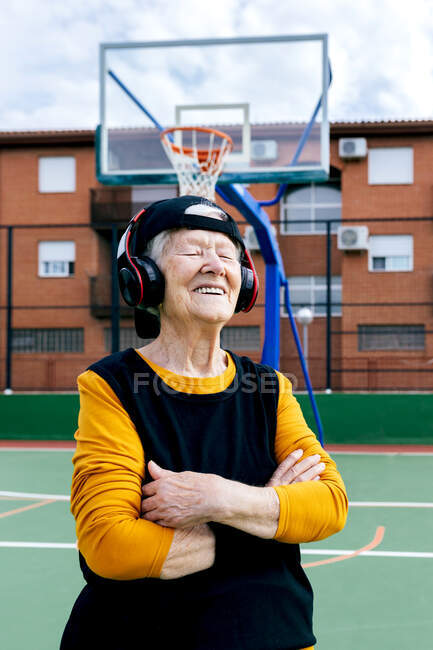 Content mature female with closed eyes in activewear listening to music with headphones while standing on public basketball court during training on street — Stock Photo