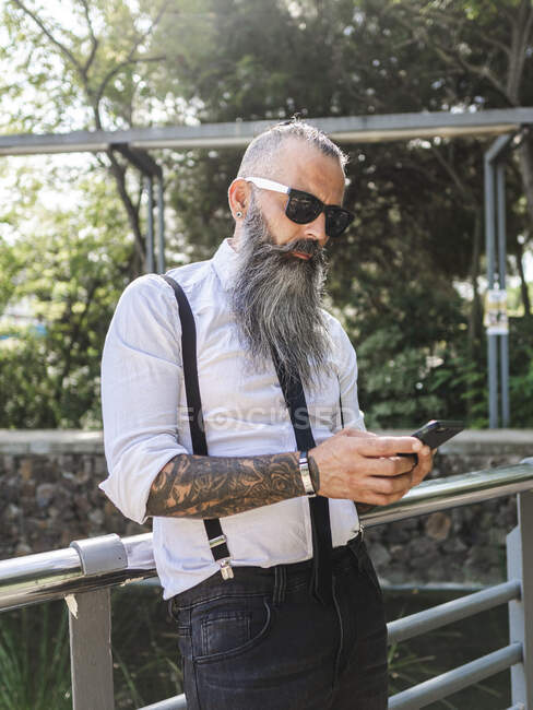 Впевнений бородатий хіпстер у стильному вбранні та текстових повідомленнях сонцезахисних окулярів на мобільному телефоні, стоячи біля металевого паркану на сонячній вулиці — стокове фото