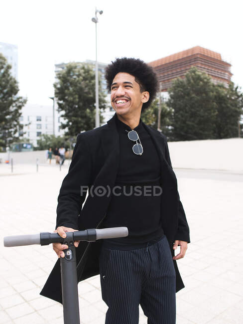 Trendiger junger Afroamerikaner mit dunklem lockigem Haar in stylischem Outfit steht auf dem Stadtplatz und schaut nach dem Rollerfahren weg — Stockfoto