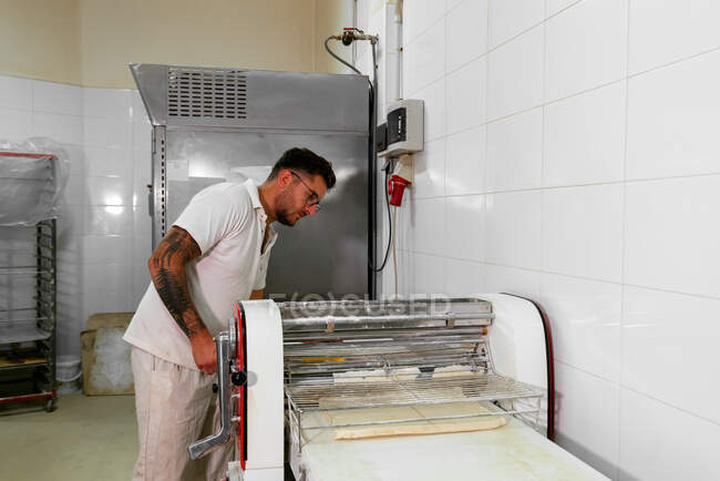 Vista lateral del chef masculino concentrado en ropa casual y anteojos de pie y trabajando en la máquina laminadora de masa mientras se prepara la pastelería en la cocina profesional - foto de stock
