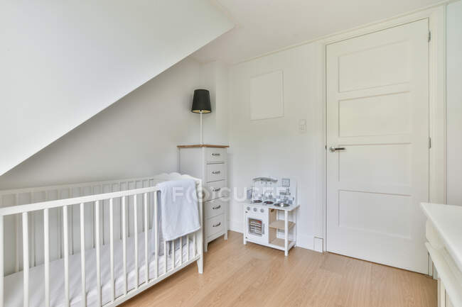 Presepe bianco con asciugamano posizionato in luce elegante camera da letto con lampada su cassettiera e giocattolo vicino alla porta chiusa — Foto stock