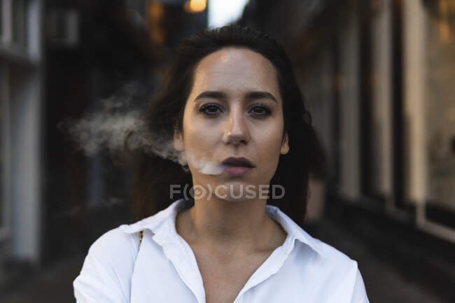 Mujer segura en camisa blanca exhalando humo mientras está de pie en la calle y mirando a la cámara - foto de stock