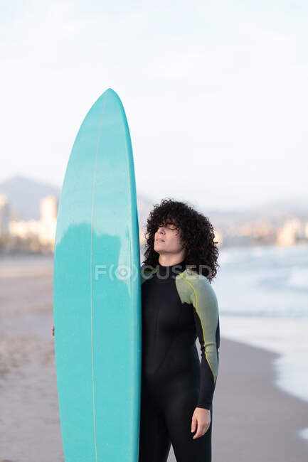 Young thoughtful female surfer in wetsuit with surfboard standing with eyes closed on seashore washed by waving sea — Stock Photo
