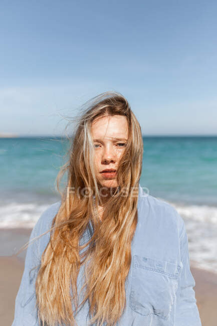Jeune femme en chemise décontractée debout sur une plage de sable près de la mer tout en regardant la caméra en été — Photo de stock