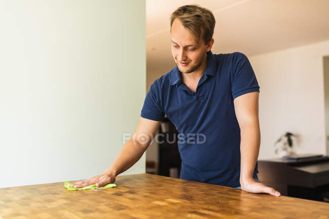Jovem macho positivo vestindo camisa de pólo azul marinho casual limpando mesa de jantar de madeira com pano amarelo — Fotografia de Stock