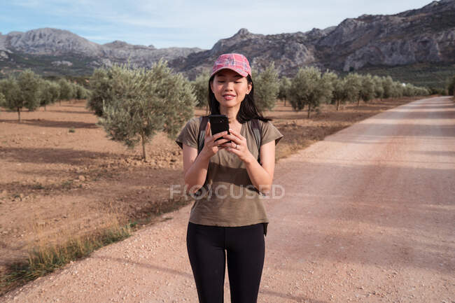 Conteúdo Asiática viajante do sexo feminino surfando celular enquanto está em pé no caminho perto da plantação com oliveiras verdes exuberantes — Fotografia de Stock