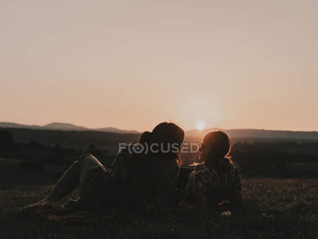 Coppia di hippie con occhiali da sole sdraiati su un prato erboso nella natura mentre osservano il tramonto tra le montagne — Foto stock