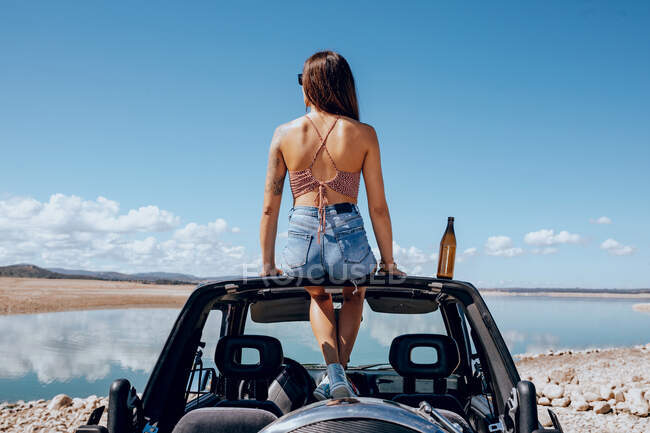 Full body of happy young female in summer outfit sitting near beer on roof of safari car on shore of river — Stock Photo