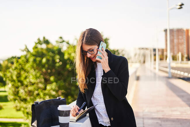 Сконцентрированная деловая женщина с каштановыми волосами в стильной одежде, стоящая возле бетонного забора с чашкой кофе и сумкой на вынос и белыми заметками в блокноте на улице в солнечный день, разговаривая по мобильному телефону — стоковое фото