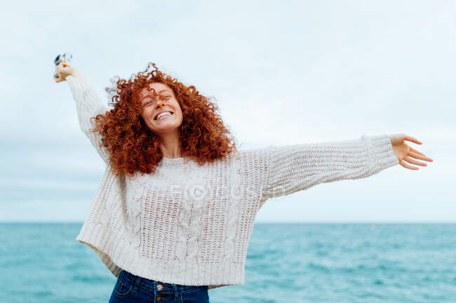 Позитивна жінка-імбир у в'язаному светрі, що стоїть з закритими очима, тримаючи ретро фотоапарат на узбережжі моря — стокове фото