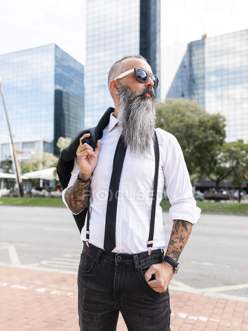 Hombre barbudo seguro con tatuaje en gafas de sol con un atuendo elegante mirando hacia otro lado mientras está parado en la calle con edificios modernos en la ciudad - foto de stock