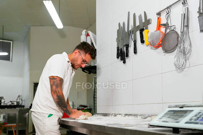 Боковой вид молодого татуированного пекаря в белой рубашке поло и очках, разминающего тесто руками, стоя у металлического прилавка на кухне современной пекарни — стоковое фото