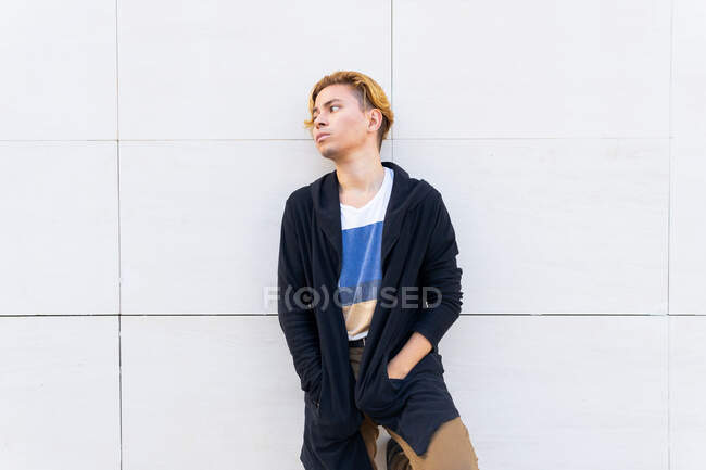 Grave giovane maschio in moderno cardigan nero guardando in lontananza con sguardo premuroso mentre in piedi vicino al muro bianco sulla strada — Foto stock