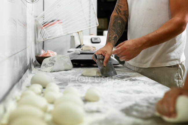 Cultivo irreconocible tatuado panadero masculino cortar piezas de masa cruda mientras se trabaja junto con su colega en la mesa de harina en la cocina - foto de stock