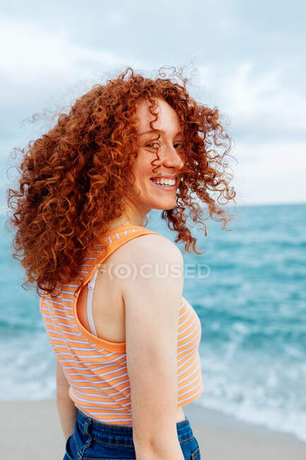 Back view of delighted female with curly long ginger hair standing looking over the shoulder to the camera against blue sea — Stock Photo