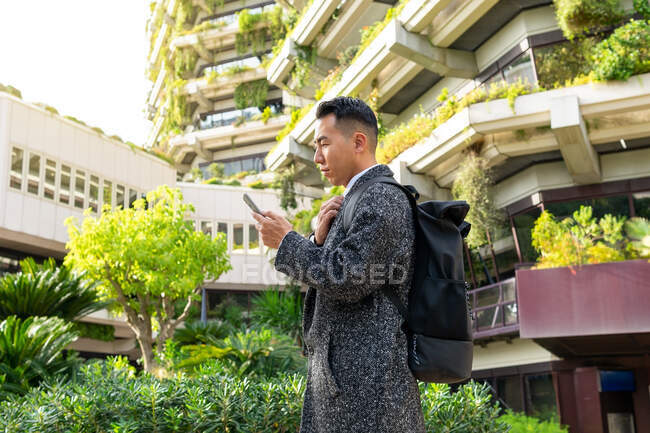 Desde abajo joven empresario étnico masculino con corbata mirando a la pantalla mientras habla por teléfono celular en la ciudad - foto de stock