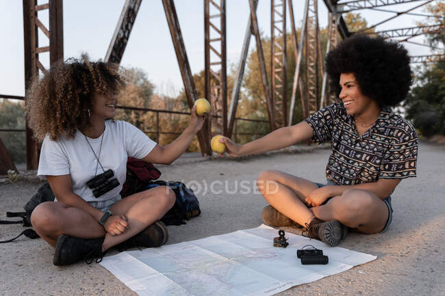 Pieno corpo di escursioniste afroamericane con mele per uno spuntino salutare seduti con le gambe incrociate e fare percorso con mappa e bussola — Foto stock