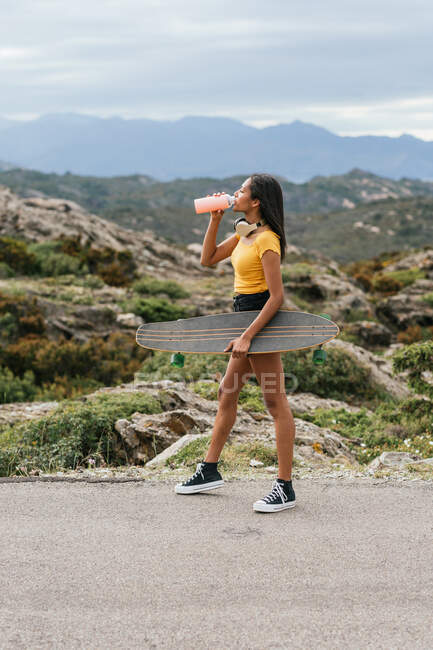 Side view of cheerful young ethnic female skateboarder with longboard standing on walkway while looking away against mountain and sea — Stock Photo