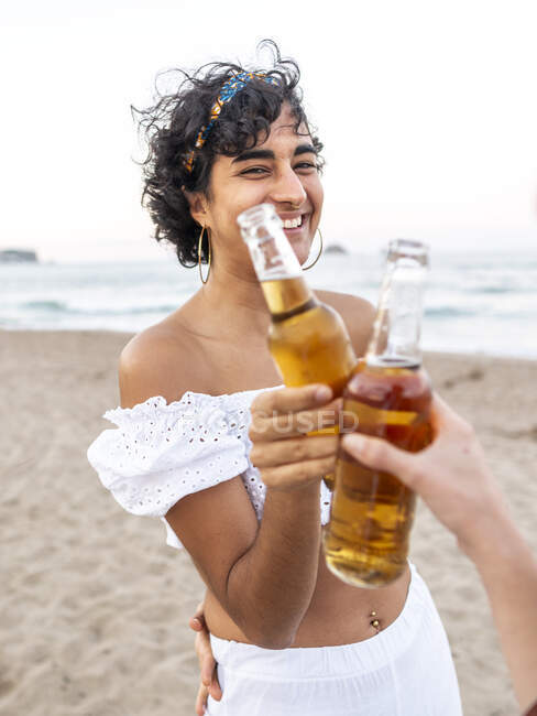 Garrafa de cerveja étnica feminina feliz com amigo da colheita enquanto desfruta da noite de verão na praia arenosa — Fotografia de Stock