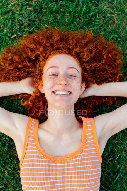 Vue du dessus de la femme aux cheveux bouclés heureux avec les mains derrière la tête relaxant sur la pelouse et regardant la caméra — Photo de stock
