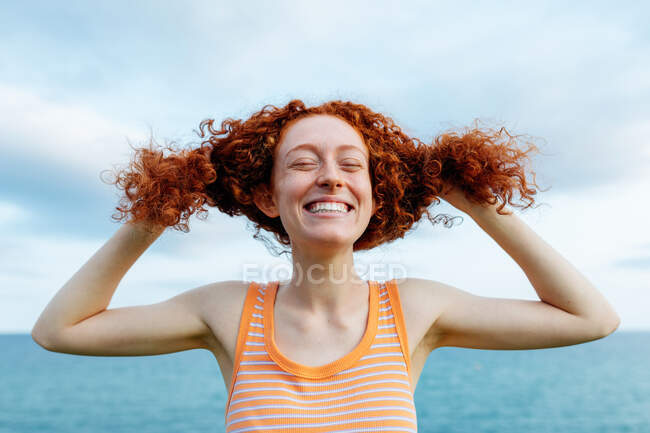 Joyful young redheaded female making childish ponytails with curly hair while enjoying freedom on seashore — Stock Photo